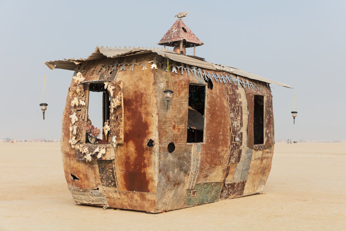 The Little Schoolhouse of Hope and Fear by: Gratis from: Vista, CA year: 2018 If all you had was the rough detritus of war, the cheap rubble of constant calamity, what would you build with it? A bunker, a church, or a school? Would you create a place of hope or fear? Of the past or for the future? Hope rising up from a violent and chaotic past, Fear transformed into building blocks: found metal, often riddled by gunre, torn by the wind and baked by the desert sun are incorporated into this replica of the traditional "little red schoolhouse." The Little Schoolhouse of Hope and Fear stands on the playa as a beacon to those who wish to teach a better way for the future, or to those ready to learn from the failures of the past.