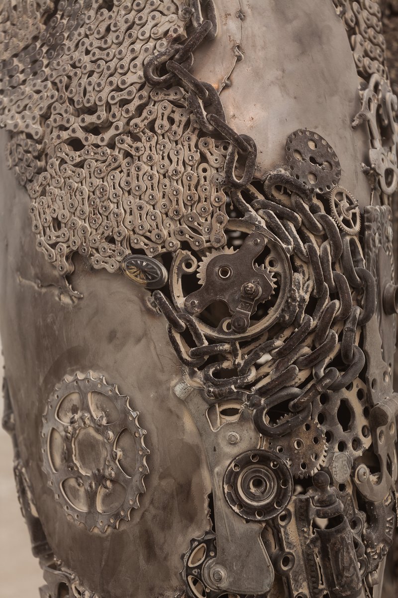 Passage by: Dana Albany from: San Francisco, CA year: 2018 Passage is an installation of a man, in a captive gesture, leisurely walking while its hand sweeps through stalks of wheat. The sculpture clearly highlights characteristics of a human form; muscle, sinuous tendon, sculpted hands, feet. Yet, it's a composite of mechanical debris and steel. Viewers may hypothesize, is this a man? is it part robot? and what is the blurry fine-line between each? The other huge component is the presence of the wheat. It symbolizes abundance, life, fertility. It is food, subsistence, of which without, the human race would be extinct. Viewers can look closer at the inherent beauty of the human form, contemplate mechanics/technology or most importantly, be reminded of our absolute reliance on nature in the end.
