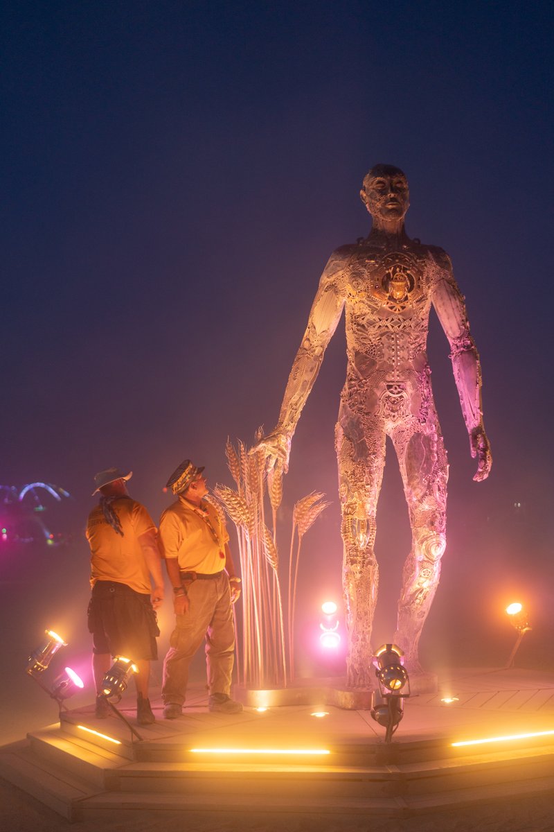 Passage by: Dana Albany from: San Francisco, CA year: 2018 Passage is an installation of a man, in a captive gesture, leisurely walking while its hand sweeps through stalks of wheat. The sculpture clearly highlights characteristics of a human form; muscle, sinuous tendon, sculpted hands, feet. Yet, it's a composite of mechanical debris and steel. Viewers may hypothesize, is this a man? is it part robot? and what is the blurry fine-line between each? The other huge component is the presence of the wheat. It symbolizes abundance, life, fertility. It is food, subsistence, of which without, the human race would be extinct. Viewers can look closer at the inherent beauty of the human form, contemplate mechanics/technology or most importantly, be reminded of our absolute reliance on nature in the end.