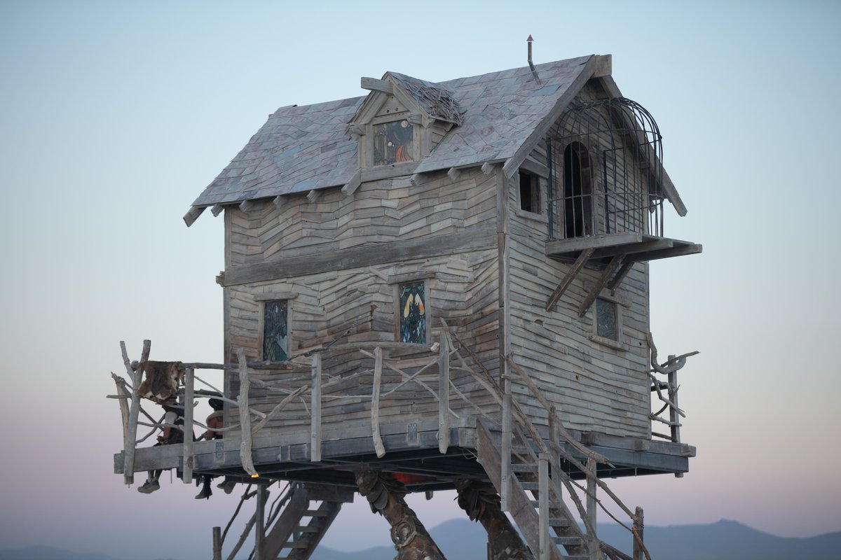 Baba Yaga's House by: Jessi Sprocket Janusee and Baba Yaga's Book Club from: Reno, NV year: 2018 The house of Baba Yaga will rise from the playa straddling it's mechanical chicken legs. The crone's house will be poised to run at the slightest provocation. Ascend her staircases to test the old witch if you dare. Are you able to walk through the veil of mortality into the space where the seer dwells? If you are strong of will, feeling bold and willing to show vulnerability she may allow you to venture within her sanctum. URL: http://jessisprocket.com/baba-yaga/