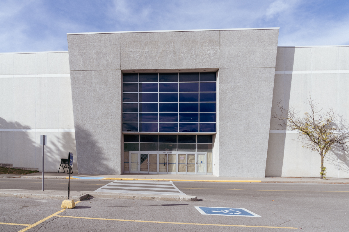 Facade of an abandoned shopping mall with boarded-up doors, tinted windows, and a deserted parking lot under a clear blue sky.